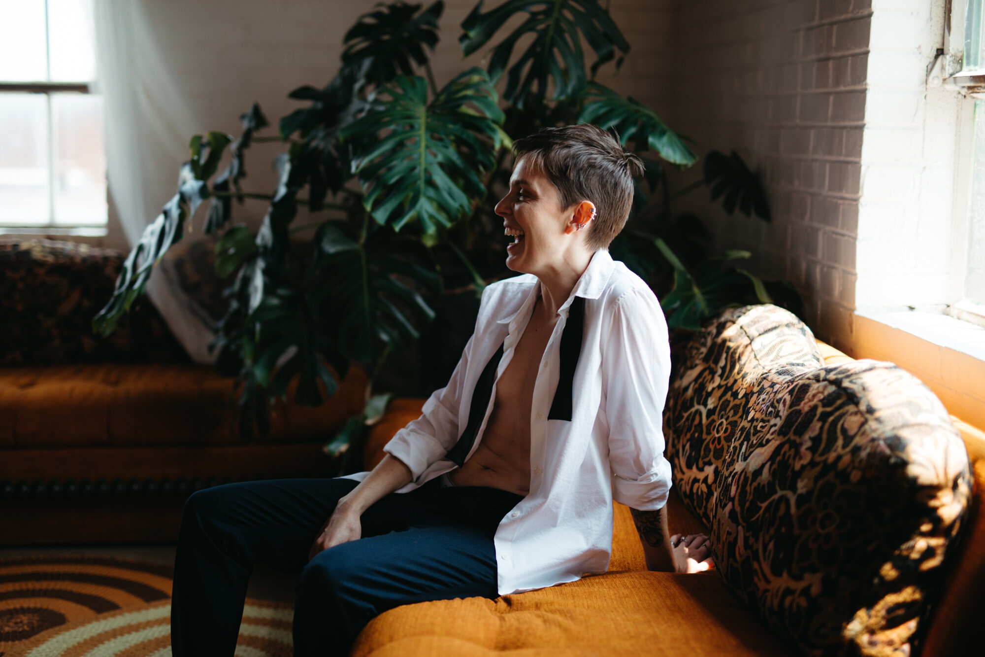 Queer person in masculine attire with shirt unbuttoned sitting on couch and laughing