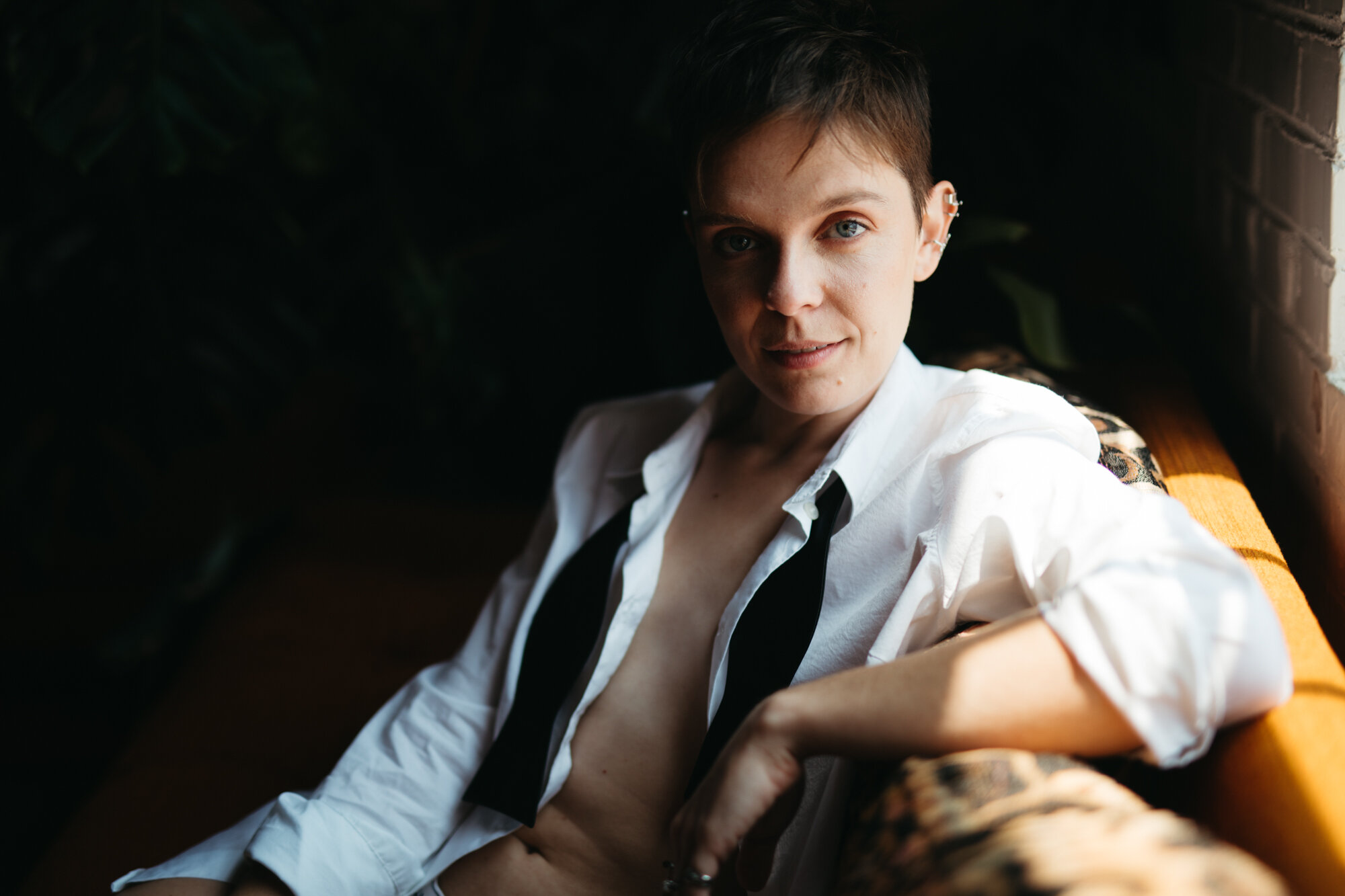Queer person in masculine attire with shirt unbuttoned sitting by window