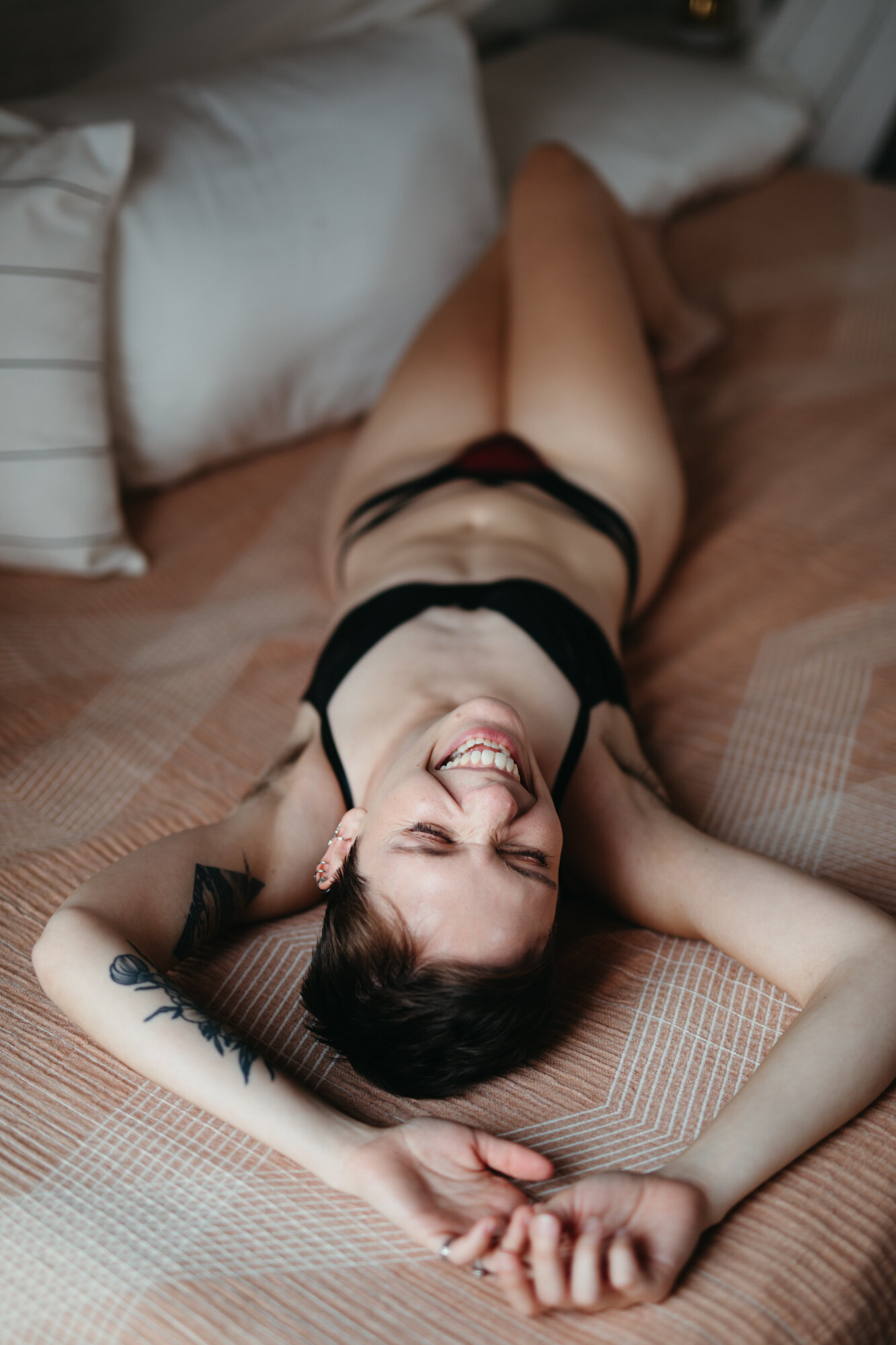 Queer boudoir photo of person laying on their back on a bed and laughing