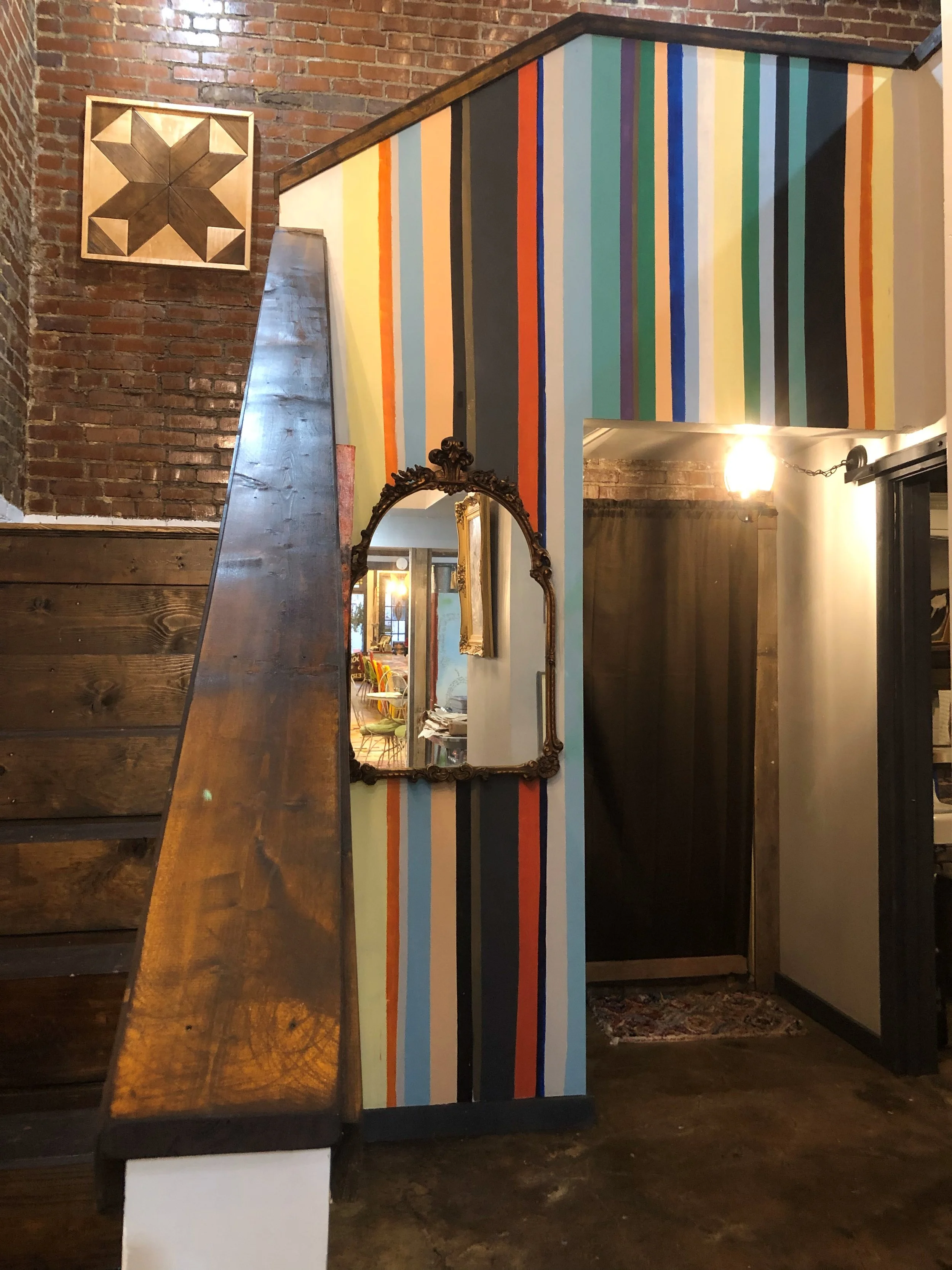 Interior view of a room with exposed brick walls, dark wood staircase, a colorful striped accent wall, and decorative elements including a framed geometric wood art piece, an ornate mirror, and warm lighting.