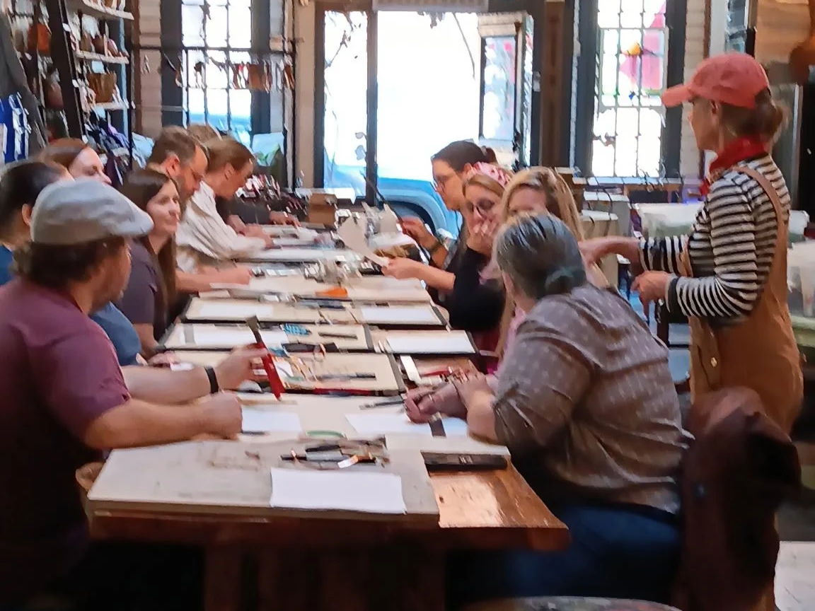 People sitting at a long wooden table in a shop or workshop, engaging in an activity involving papers and small objects, with shelves filled with items in the background and a person standing and interacting with others.