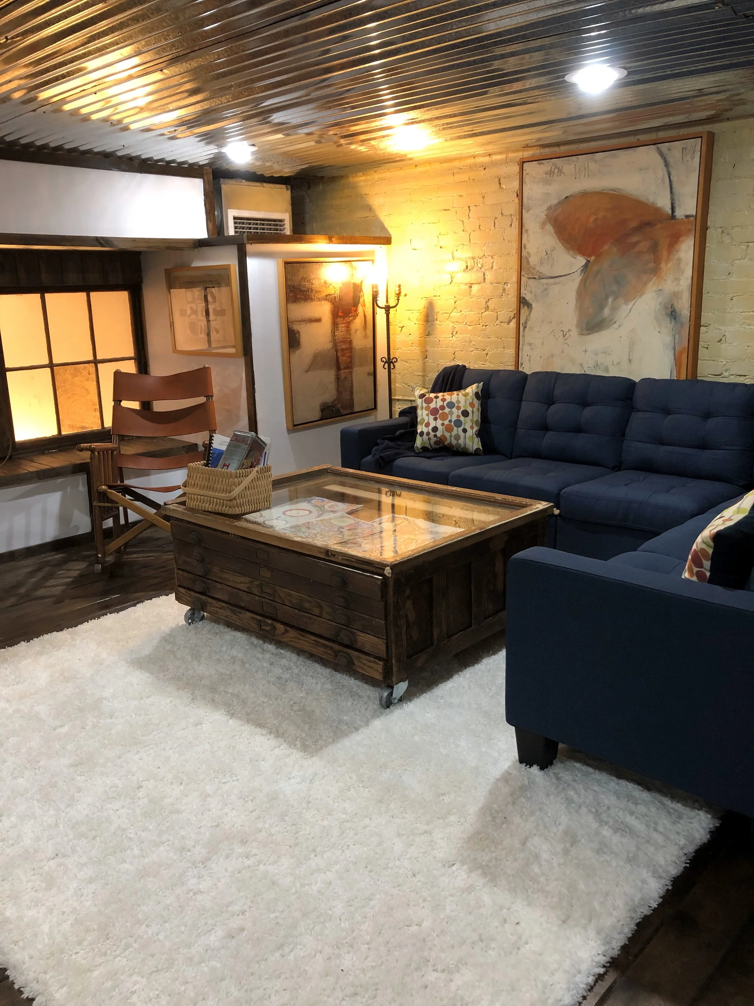 Living room with a navy blue sectional sofa, a wooden coffee table on a white shag rug, a window with black frames, abstract paintings on the brick wall, and a black standing lamp.