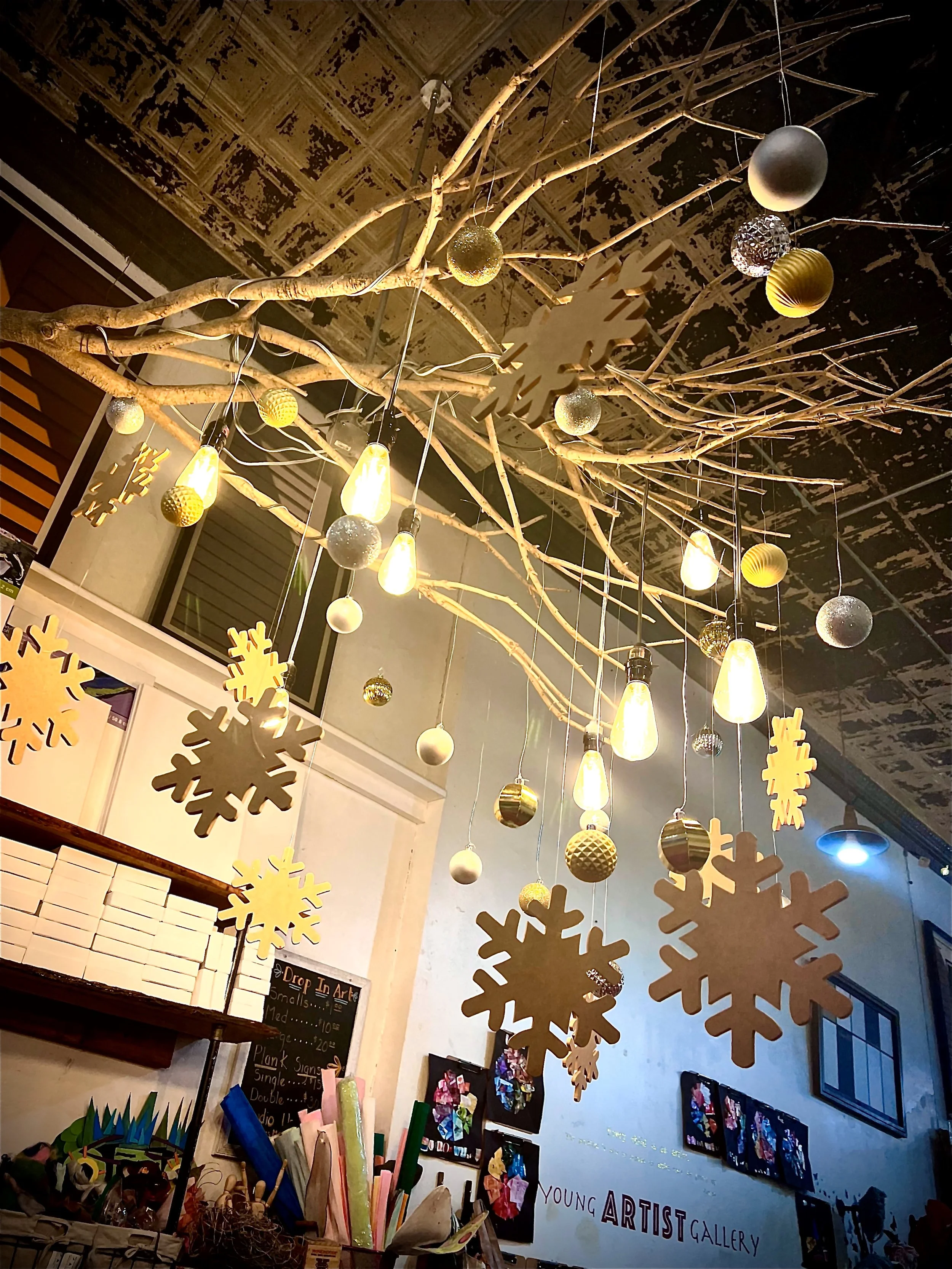 Decorative Christmas holiday display featuring hanging golden and silver balls, snowflake ornaments, and warm hanging lights, with an art gallery or shop interior background.