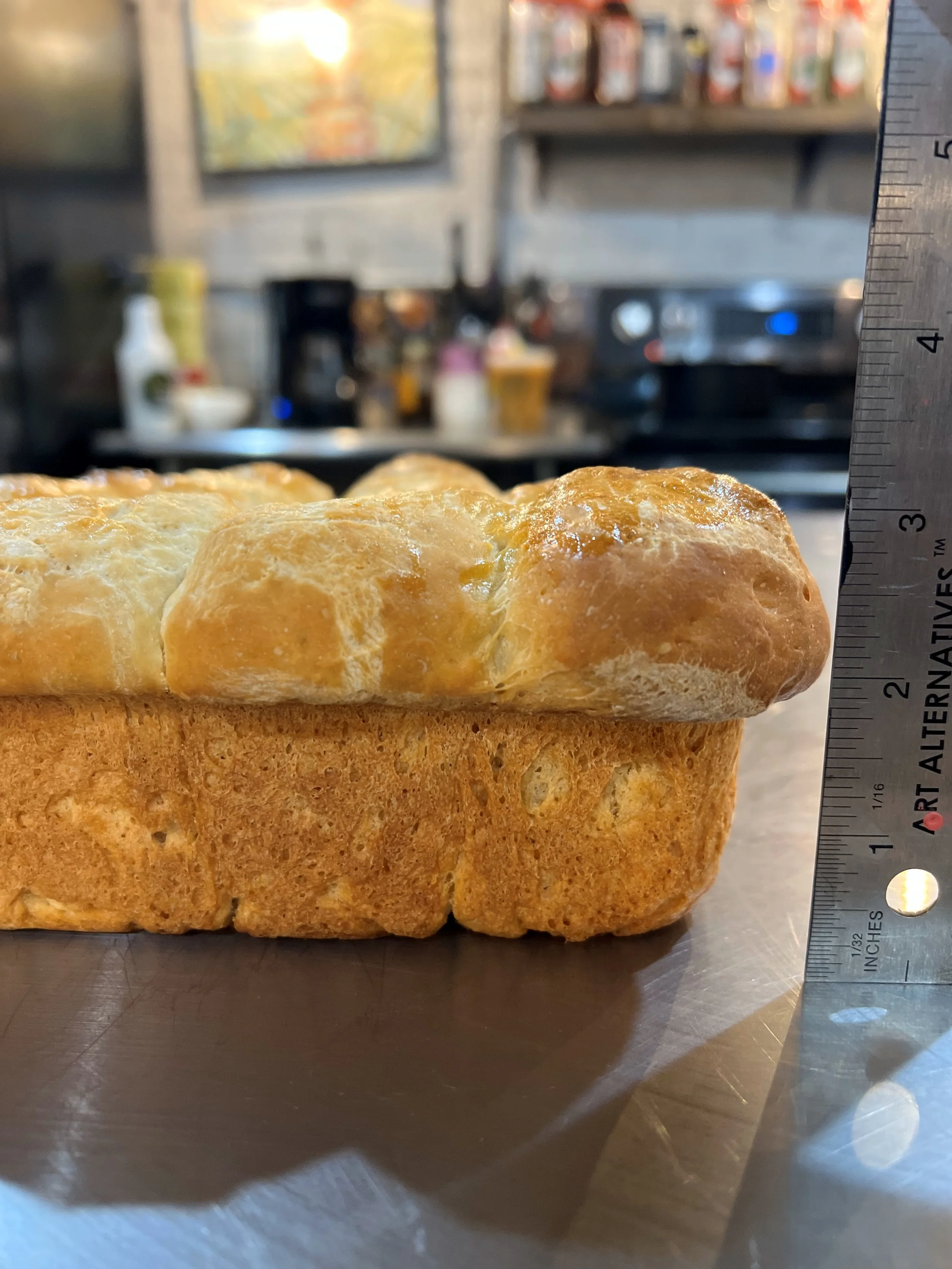 A loaf of bread on a metal countertop with a ruler standing next to it, showing its height. The background is a blurry kitchen with various items on shelves.