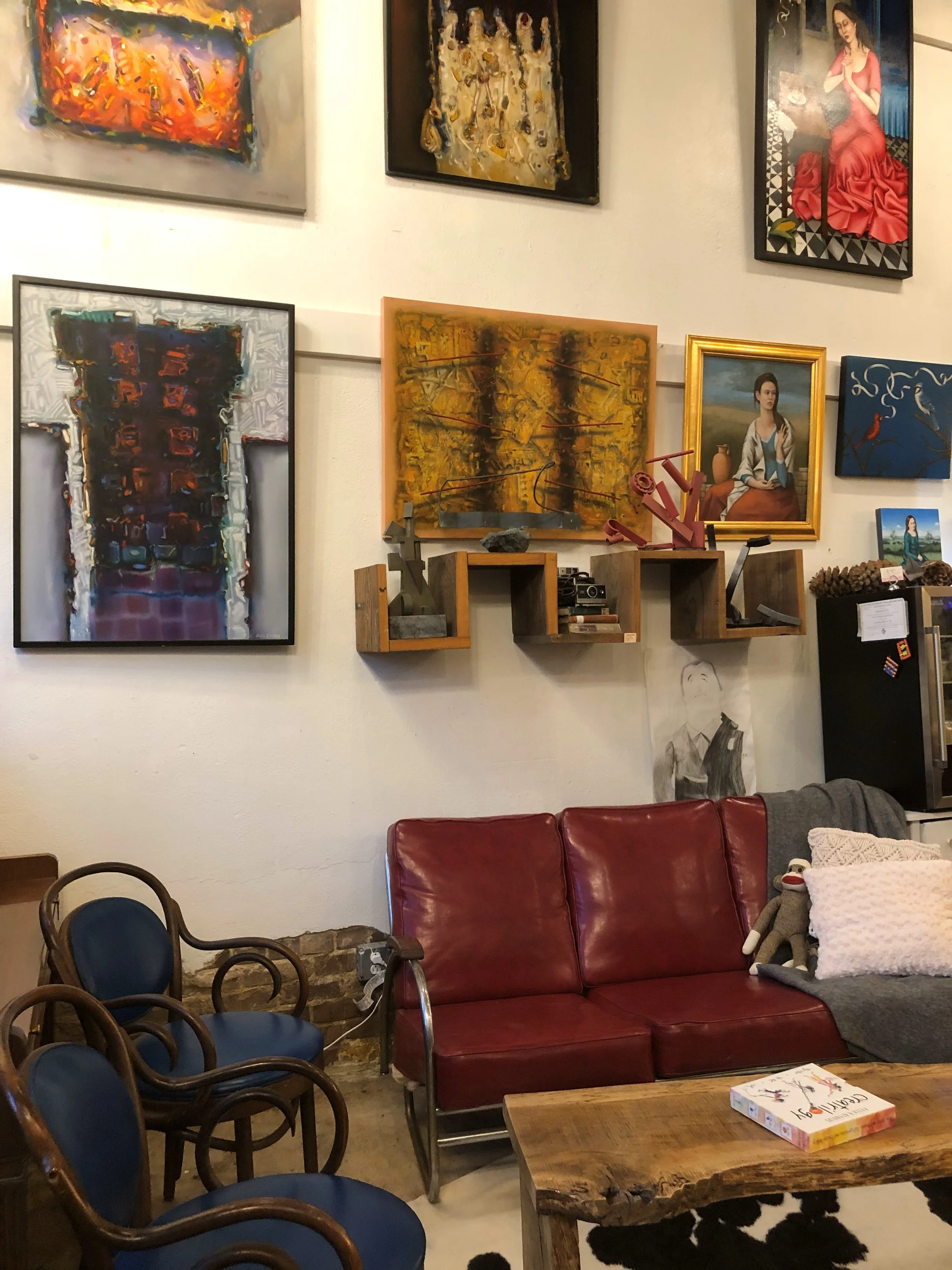 A cozy room with a collection of colorful paintings on the wall, a red leather couch, a rustic wooden table with a book on it, and several chairs in the foreground. There is a pillow and a stuffed monkey on the couch, and artwork featuring a woman in