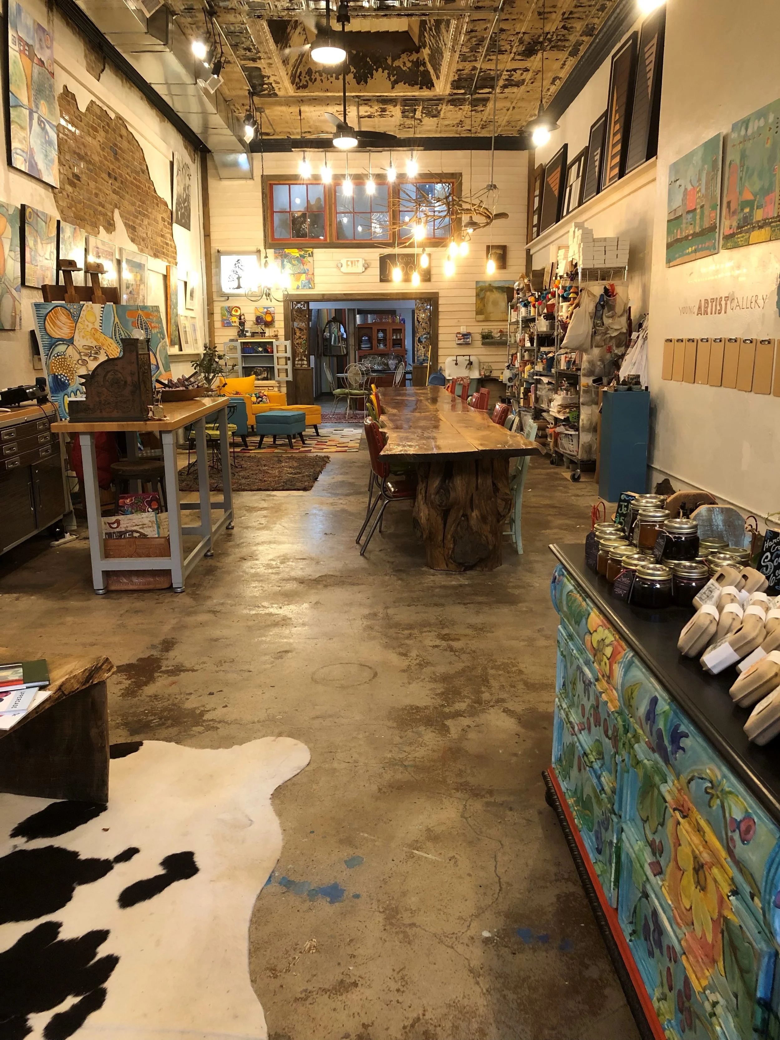 Interior of an art studio or gallery featuring colorful paintings on walls and shelves, a large wooden table with chairs, a cowhide rug, and various art supplies and displays.