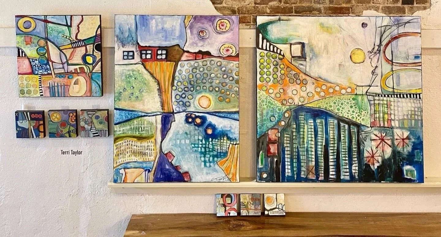 A collection of abstract paintings displayed on a wall, with a smaller set of paintings beneath them on a wooden surface. The artwork features vibrant colors, geometric shapes, and patterns, created in an abstract style.