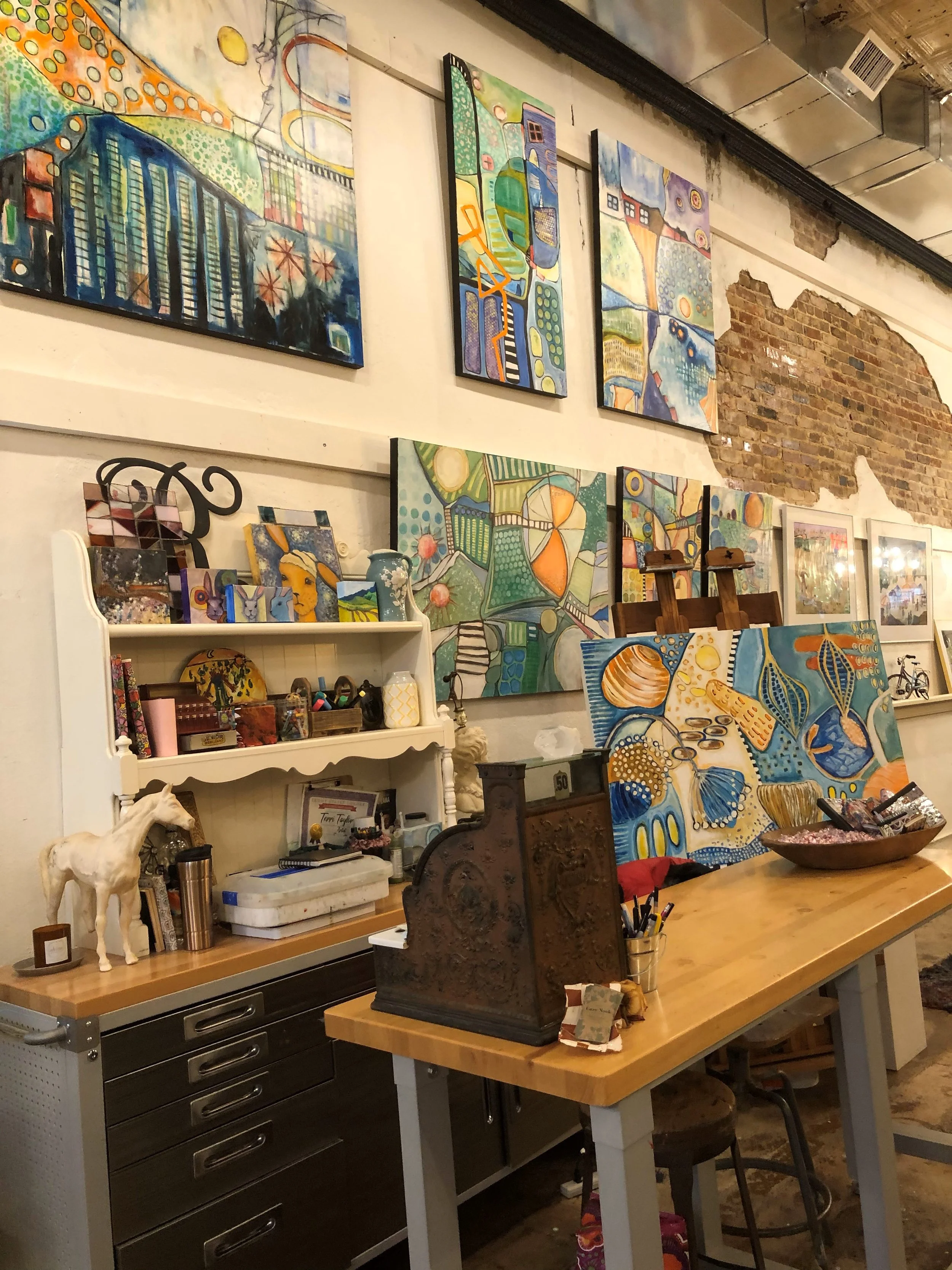 An art studio with a long wooden table, colorful abstract paintings on the wall, a white shelf with books and decorative items, and a decorative horse sculpture.