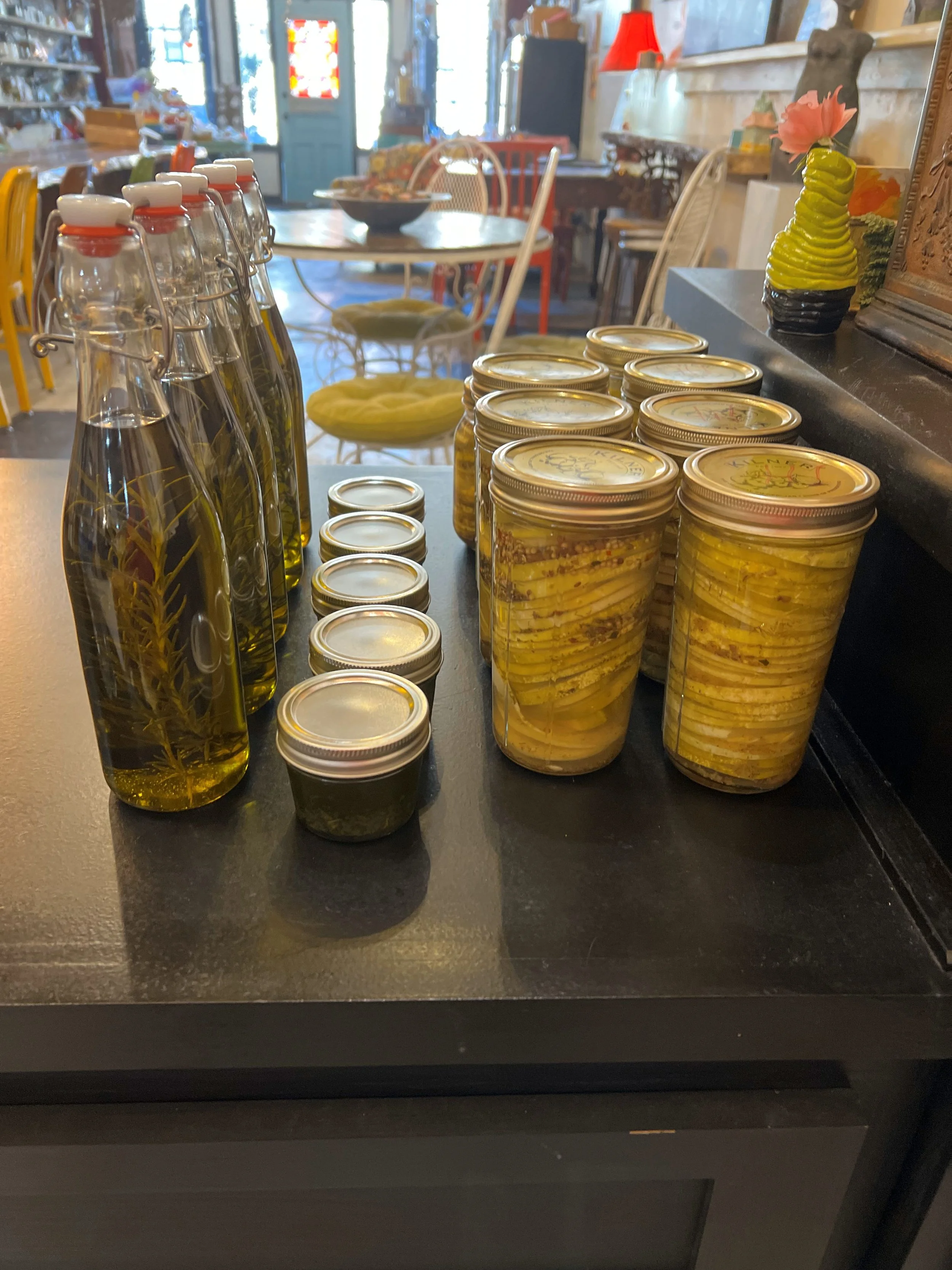 Display of bottles and jars with olive oil and preserved food on a kitchen or cafe counter, surrounded by various furniture and decorative items.