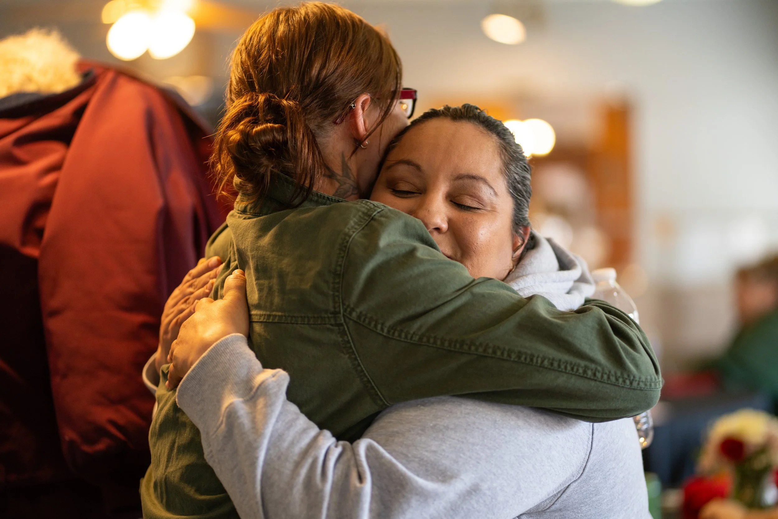 Two women exchange items at an outdoor event, with trees and people in the background.