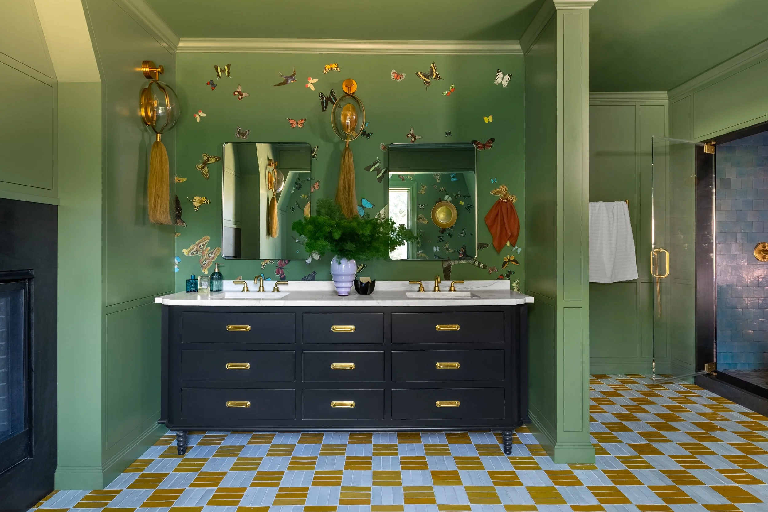Bathroom with green walls decorated with colorful butterfly and bird stickers, black vanity with gold handles, white marble countertop, two mirrors, and a plant in a purple vase.