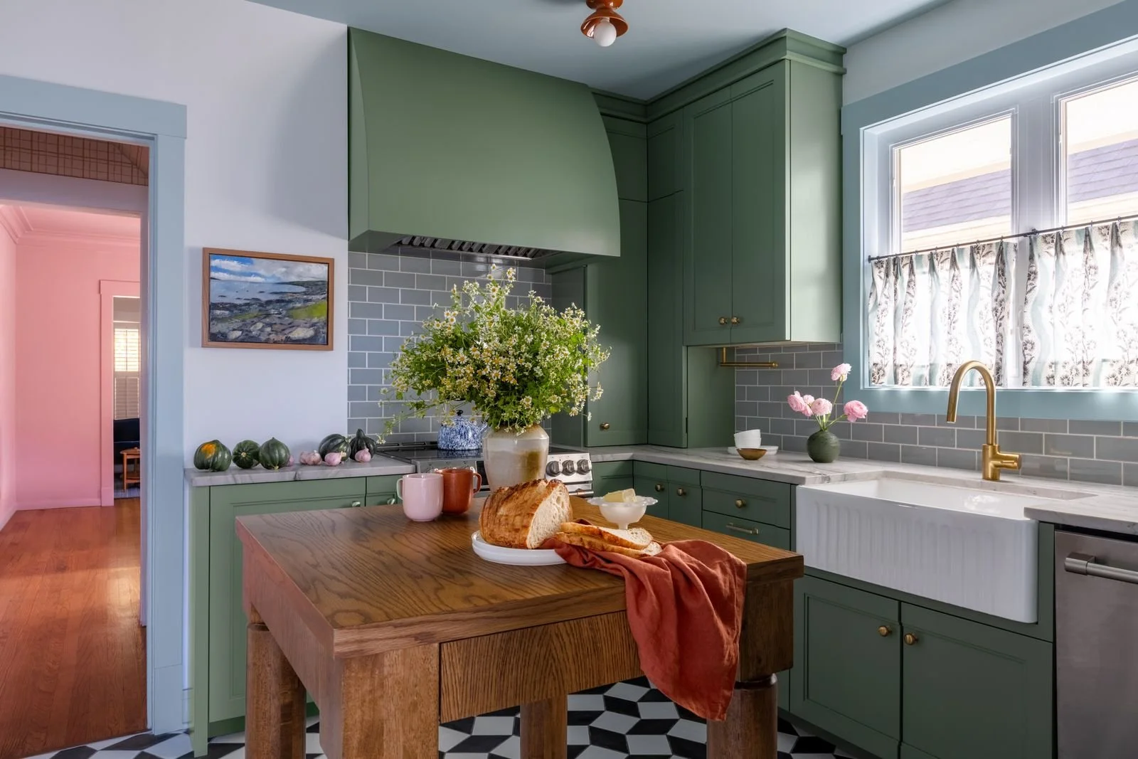 Kitchen with green cabinets, white farmhouse sink, gold faucet, window with floral curtains, wooden island with bread and breakfast items, floral arrangements, and decorative gourds.