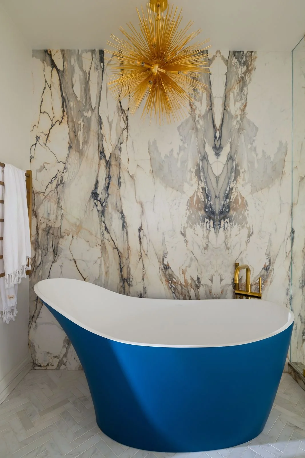 A modern bathroom with a blue freestanding bathtub, gold faucet, and a marble wall with gray, white, and black veining. There is a gold chandelier on the ceiling and a white towel hanging on a wooden rack.