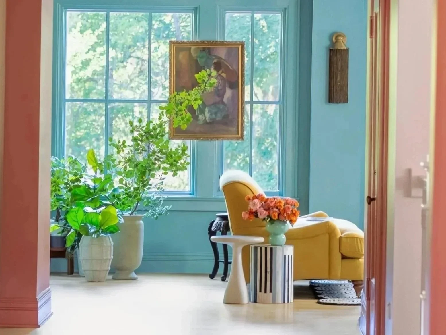 A cozy living room with large blue-framed windows, a yellow armchair, a small white side table with a vase of pink flowers, large potted green plants, and framed artwork on the wall.