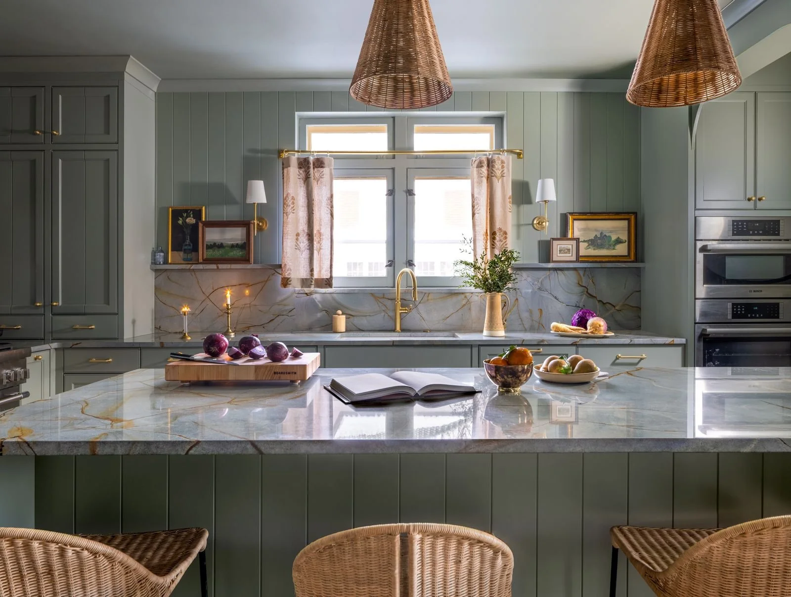 Modern kitchen with green cabinetry, marble countertops, and a window with floral curtains. Decor includes candles, framed art, potted plant, and bowls of fruit.