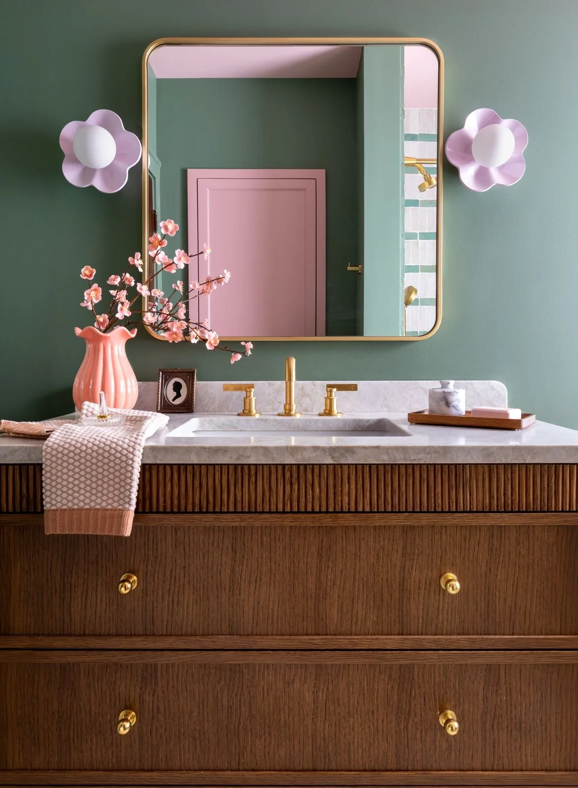 Bathroom vanity with a marble countertop, gold faucet, pink flower vase with cherry blossoms, decorative framed portrait, and pink flower-shaped wall lights reflected in a mirror with a gold frame. The vanity has a wooden finish with gold knobs.