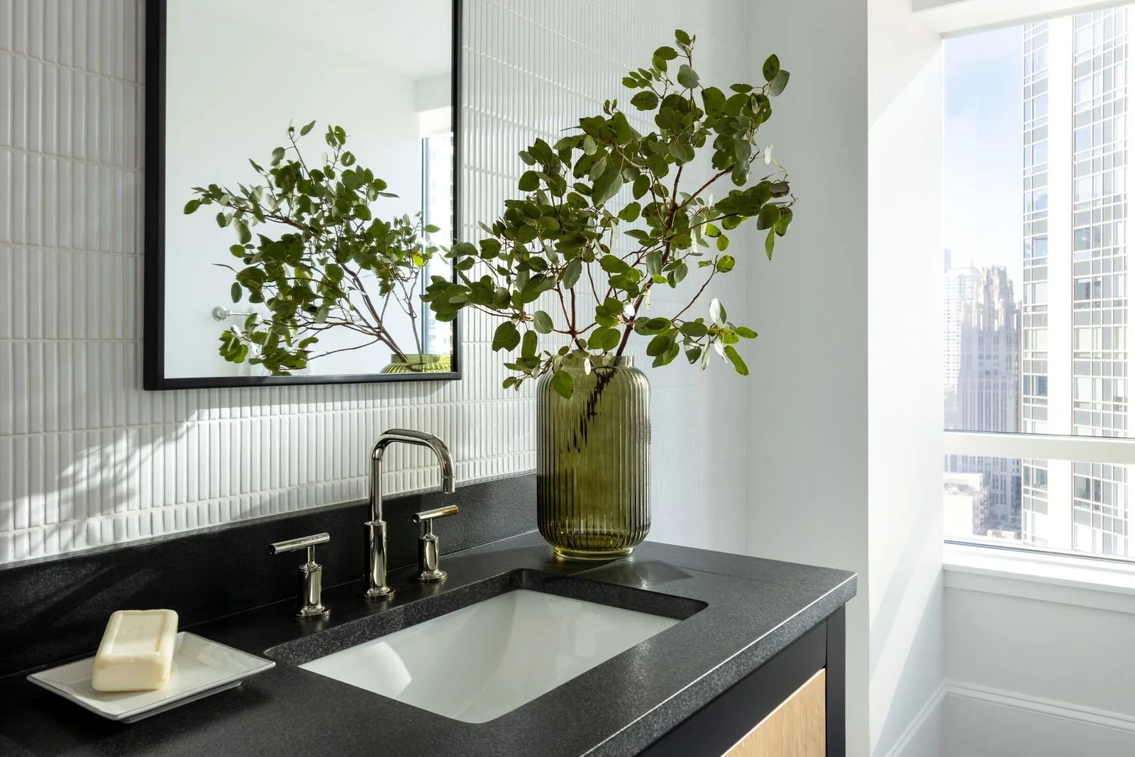 Modern bathroom with black countertop, white sink, silver faucet, large green leafy plant in a ribbed glass vase, mirror reflecting part of the plant, white textured tile backsplash, and a window with city view.