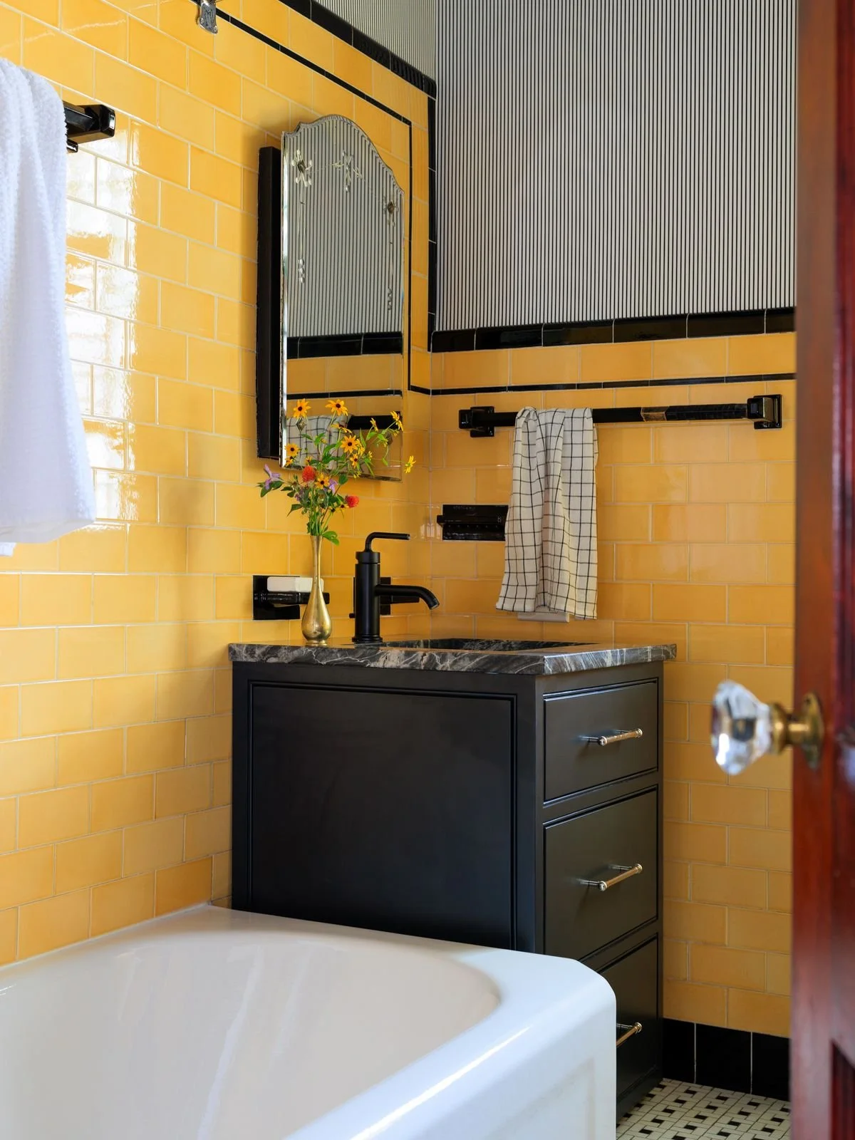 A bathroom with yellow tiled walls, a black vanity with a marble countertop, a black faucet, a mirror, and a vase with colorful flowers. A towel is hanging on a black towel bar.