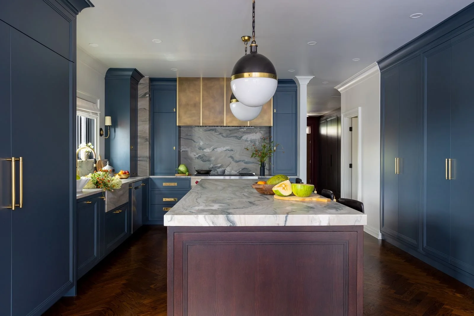 Modern kitchen with blue cabinets, white marble countertops, a marble backsplash, and a central island with a dark wood base. There is a black and gold pendant light hanging above the island. The kitchen has fruit on the counters and a window with a 