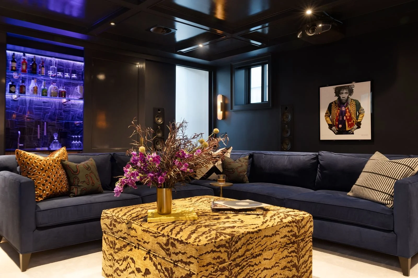 A modern living room with a dark wall, navy sectional sofa decorated with patterned and solid pillows, a tiger-print ottoman with a vase of pink and yellow flowers on top, and a small side table with a glass of drink. There is a framed black-and-whit