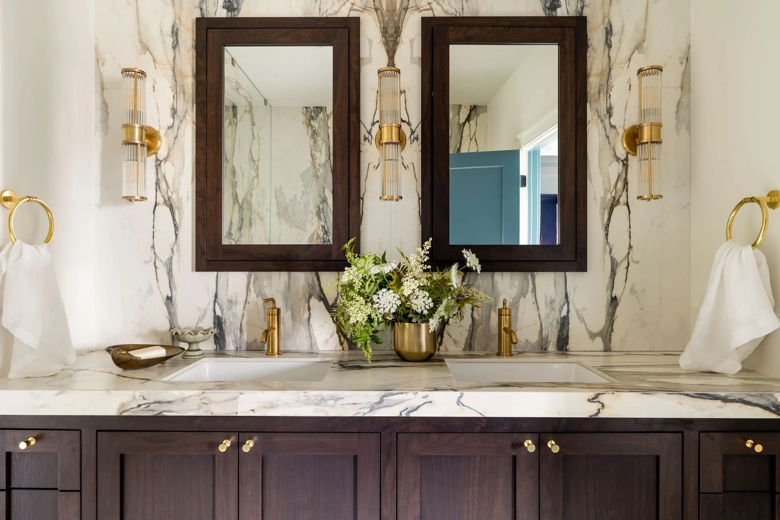 A luxurious bathroom double vanity with a marble countertop, two sinks, and wooden cabinets. There are two large mirrors with dark wooden frames above each sink, with gold fixtures and wall-mounted light fixtures. A vase with white flowers sits in th