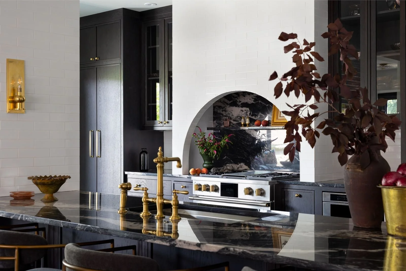 A modern kitchen with black cabinets, white brick walls, and a marble island counter with a gold faucet. There is a black and gold stove with a decorative black marble backsplash and a large arched opening. Decor includes a vase with dark red leaves,