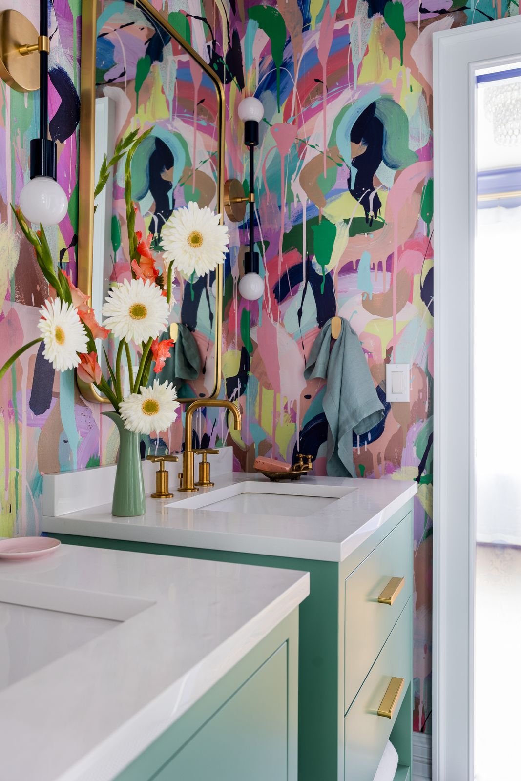 A bathroom with colorful abstract wall art, a mint green vanity with gold hardware, a white countertop with two sinks, a gold faucet, a mirror, a vase with white flowers and pink accents, and wall-mounted light fixtures with globe bulbs.