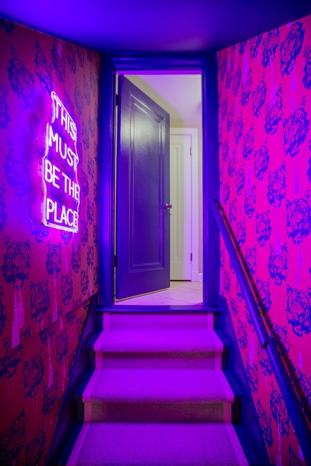 A staircase with three steps illuminated by purple lighting, leading up to a partially open dark gray door. The walls are covered in pink wallpaper with a pattern of blue tiger faces. A neon sign on the left wall reads, 'This must be the place' in wh