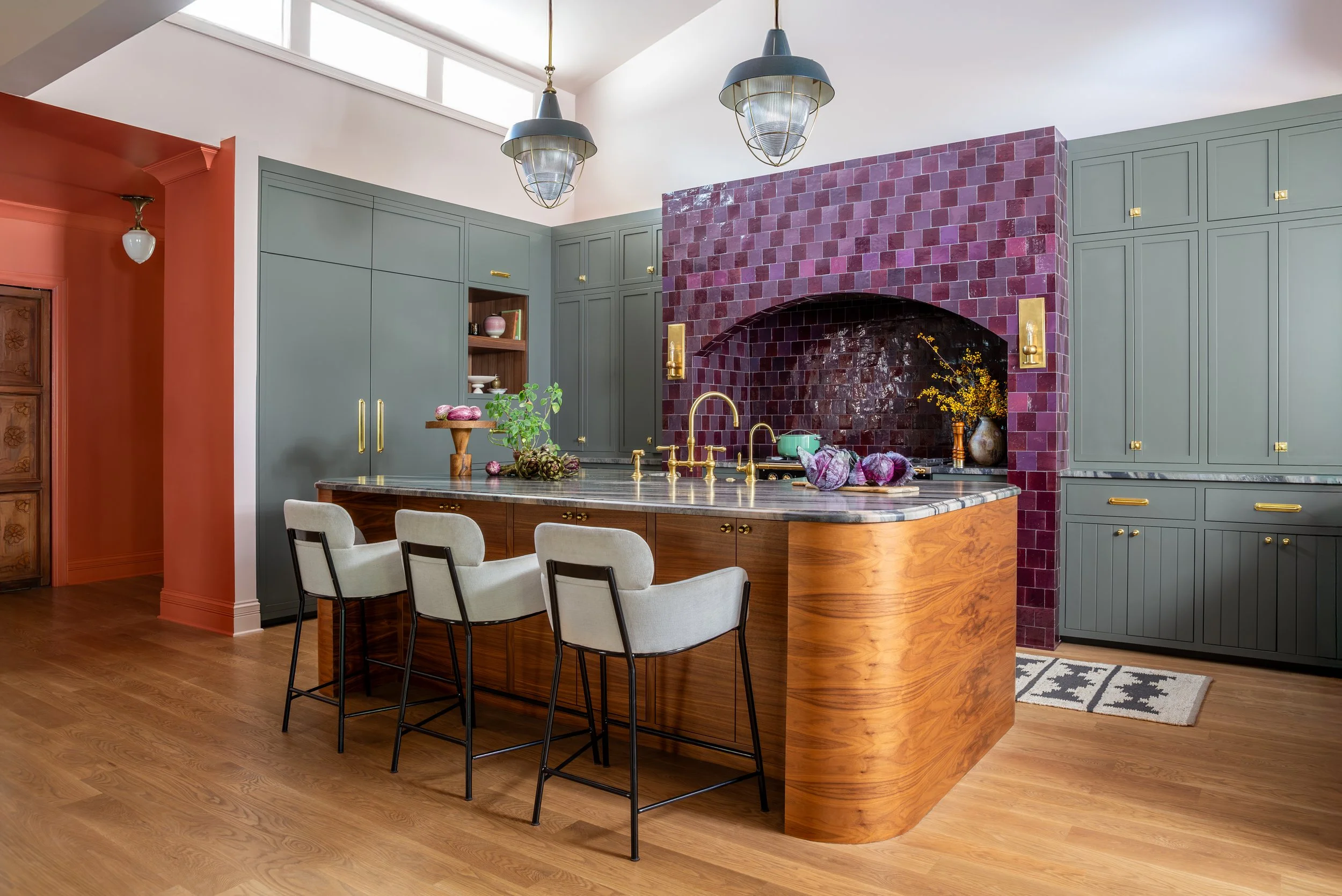 Modern kitchen with green cabinets, red tiled fireplace, wooden island with marble top, and four cream-colored chairs. Pendant lights hang from the ceiling, and there are decorative items like a vase with yellow flowers and purple vegetables on the i