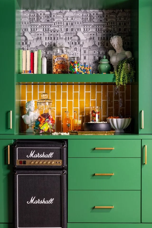 Green kitchen cabinet with open shelves displaying books, a white bust sculpture, jars, candies, and decorative items. A black Marshall guitar amplifier and speaker are below the shelves.