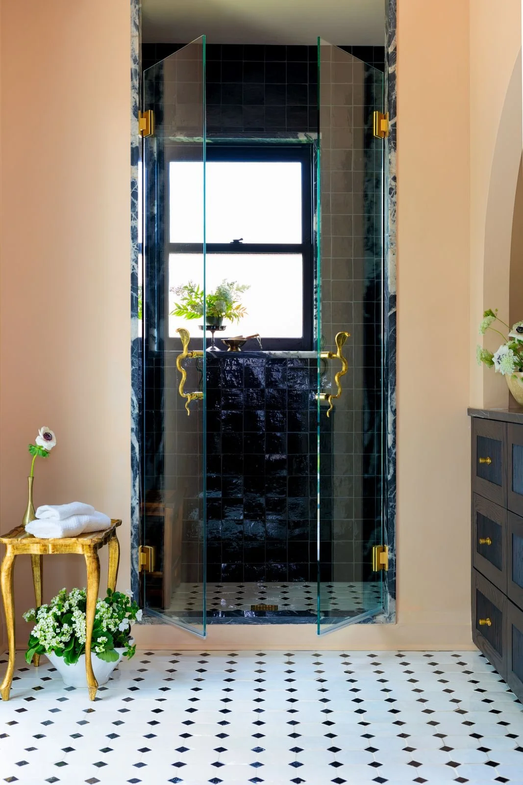 A luxurious bathroom with a black tiled shower featuring gold snake-shaped handles and a window with a potted plant on the windowsill. Adjacent is a light-colored wall, with a small striped side table holding a flower vase and towels, and a dark wood