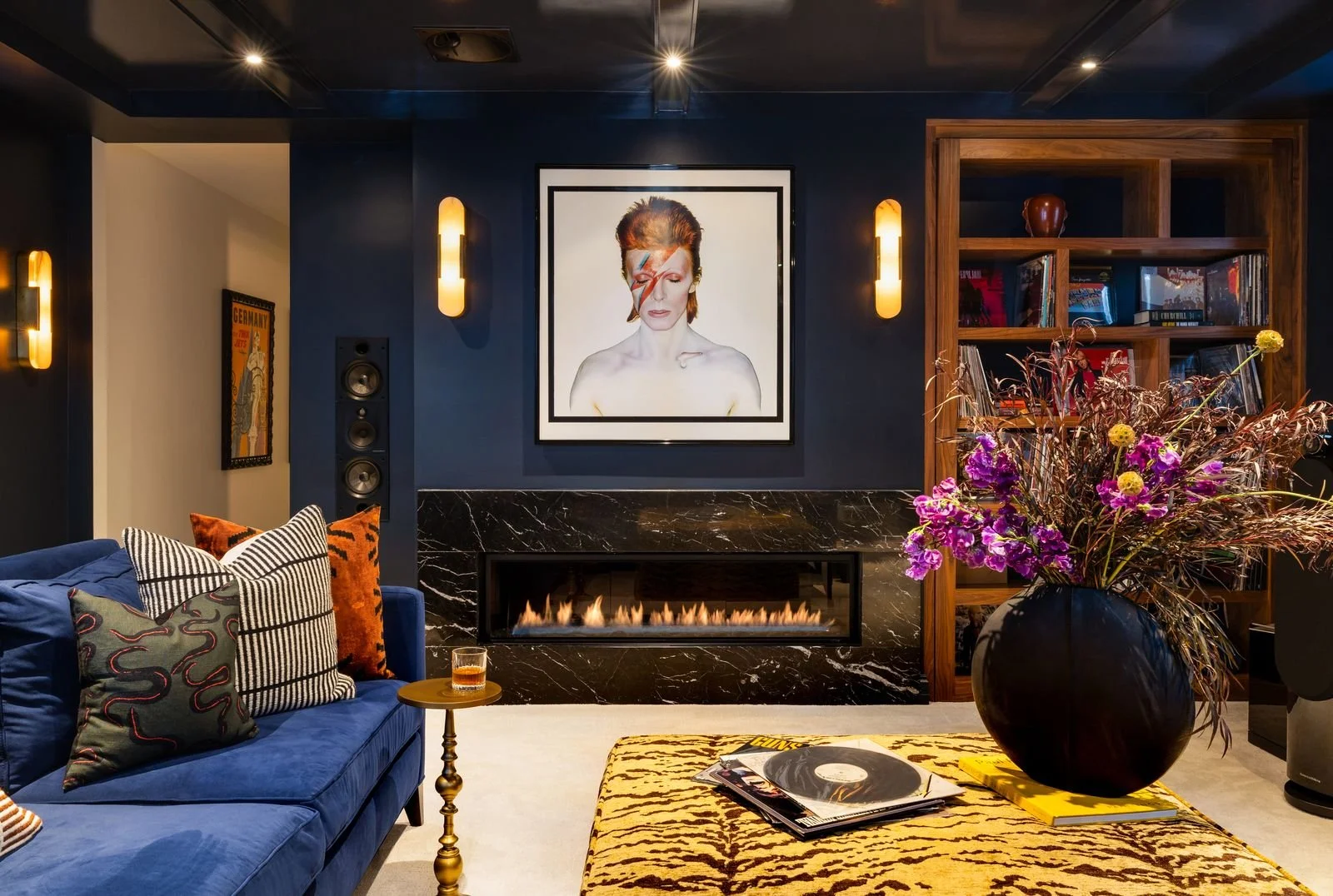 Living room with a dark blue wall, a fireplace with a black marble surround, and a framed portrait of David Bowie with a lightning bolt makeup. There is a blue velvet sofa with decorative pillows, a small gold side table with a drink, a large black v