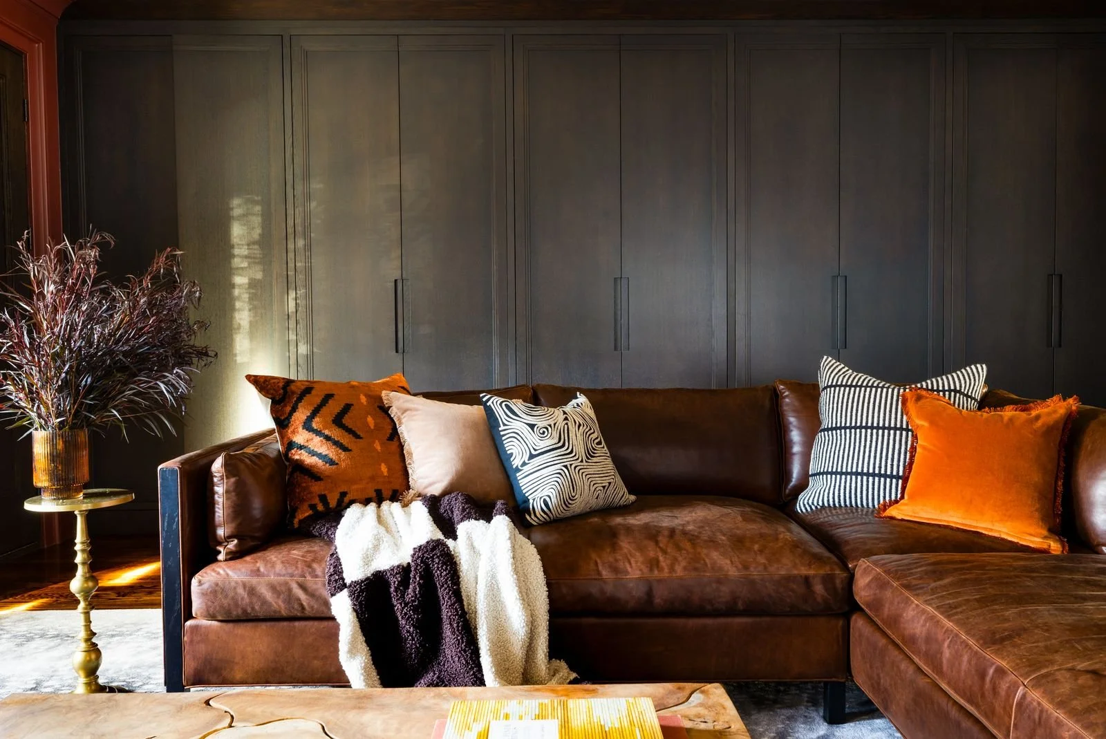 Living room with leather sectional sofa decorated with patterned and solid colored throw pillows, a cozy blanket, a side table with a tall vase of purple foliage, and built-in dark gray cabinets on the wall.