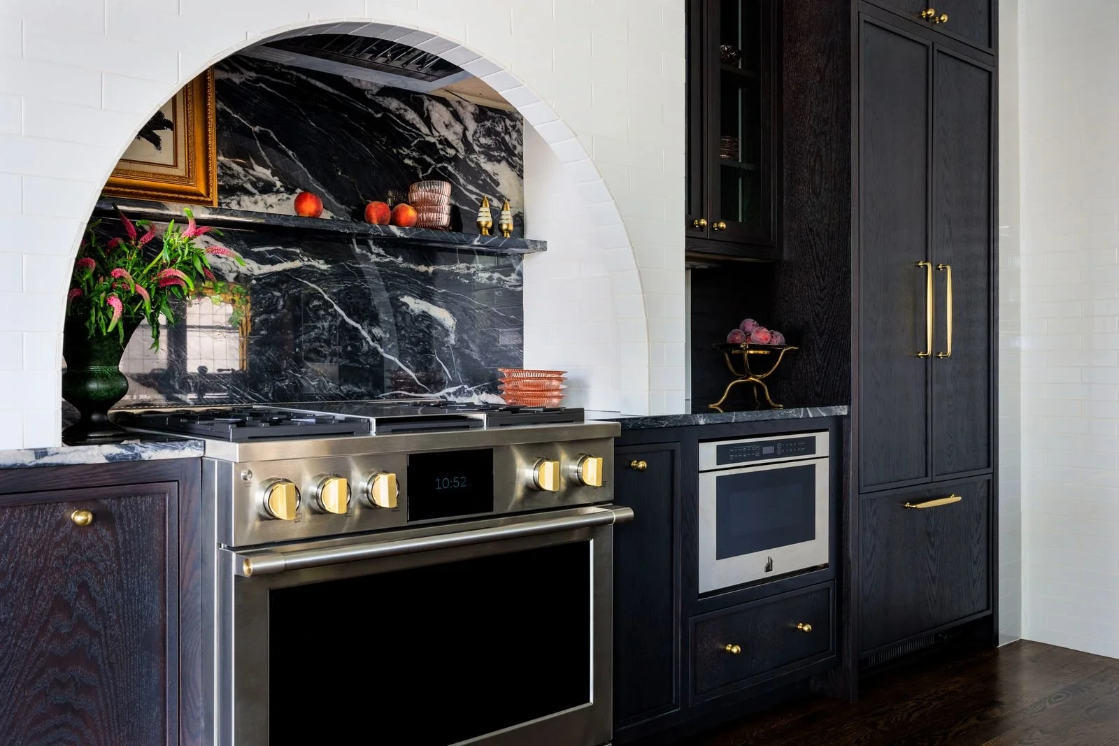 Modern kitchen with black cabinets, black marble backsplash, stainless steel oven with gold knobs, built-in microwave, and decorative items including a vase with pink flowers, apples, and purple grapes.