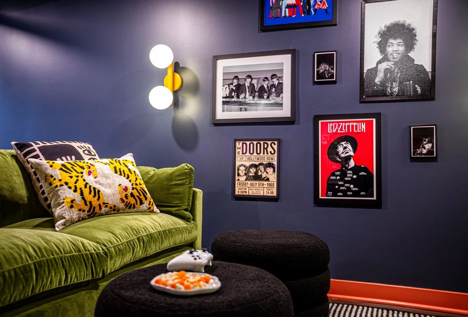 A cozy living room corner with a green velvet sofa, decorative pillows with tiger design, a black pouf, and wall art featuring famous musicians and band posters, including Led Zeppelin and The Doors, on a dark blue wall. There is a modern yellow wall