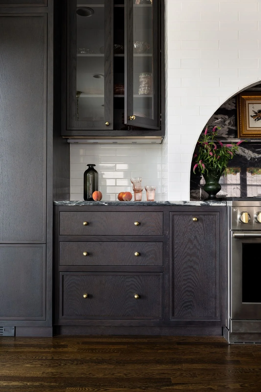 Kitchen with dark wood cabinets, peaches, pink glasses, a black vase, flowers, and a framed picture.