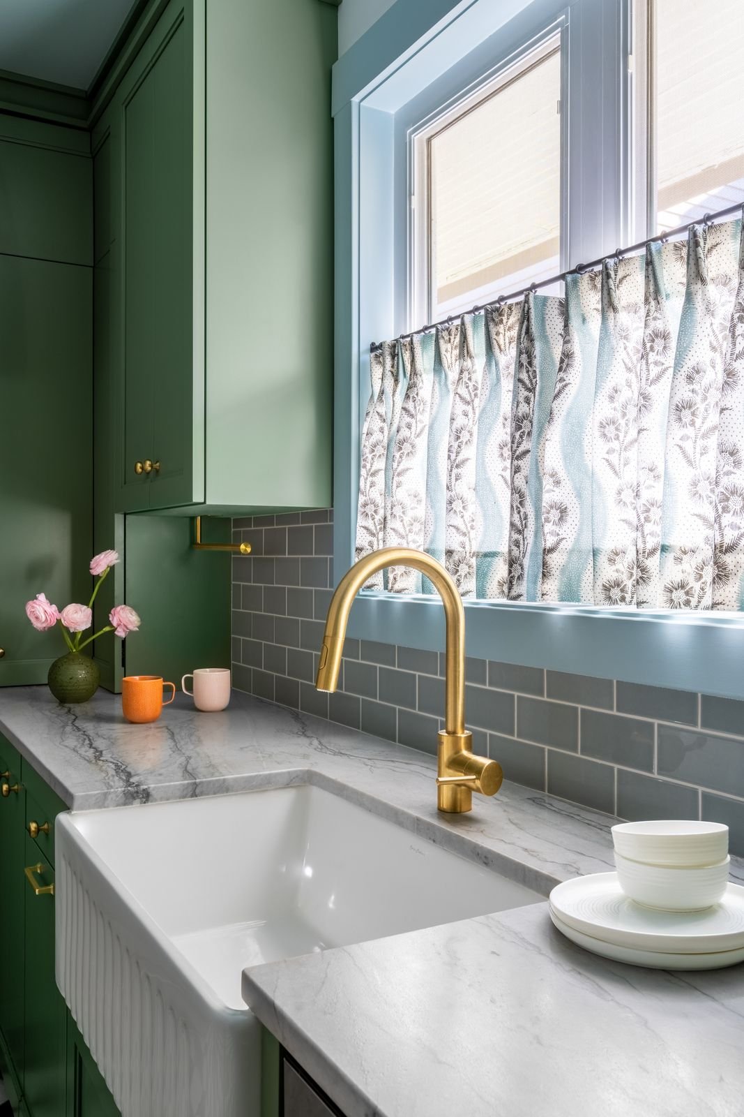 Kitchen with green cabinets, a marble countertop, a white farmhouse sink, gold faucet, gray subway tile backsplash, window with patterned curtains, and pink flowers in a green vase.