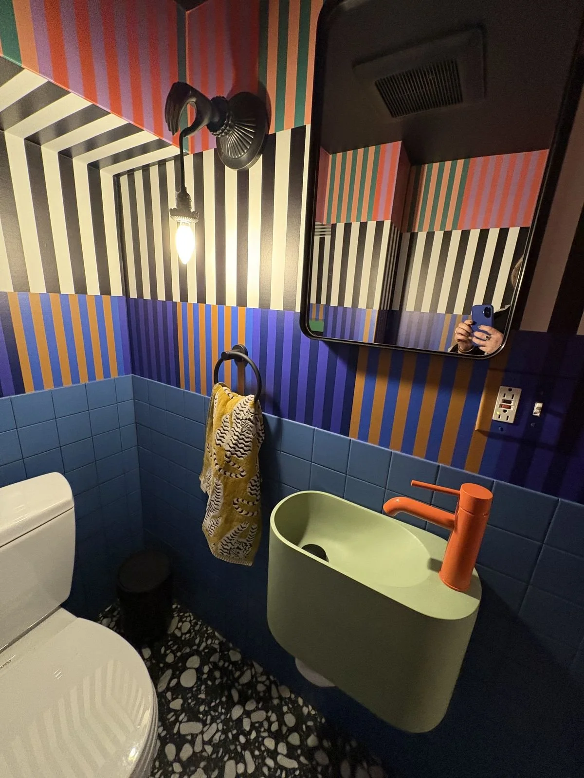 A colorful bathroom with geometric striped wallpaper in black, white, purple, teal, orange, and red. Features include a black flamingo-shaped wall light, a rectangular mirror, a yellow hand-washing sink with an orange faucet, a yellow towel with a bl