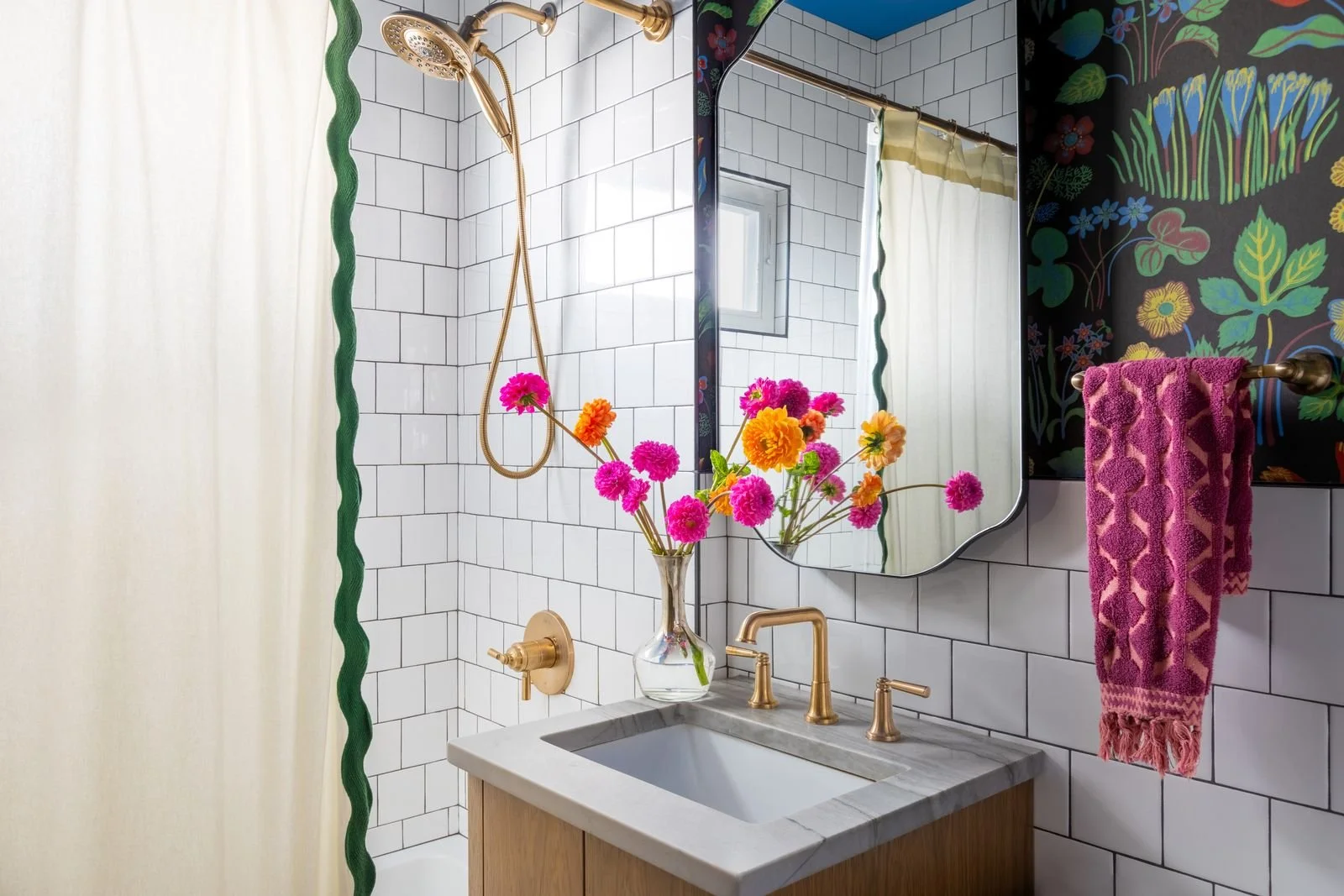 Bathroom vanity with a mirror, vase of pink and orange flowers, gold faucet, pink towel, white subway tiles, a shower with a gold showerhead and a green wavy border on the curtain, and a black floral wallpaper.