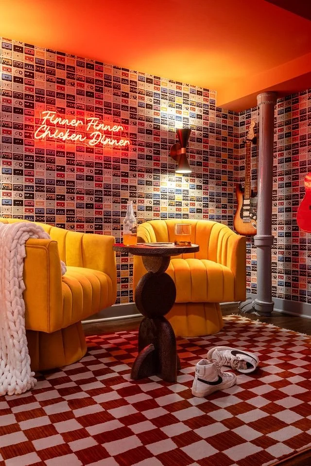 A cozy seating area with two yellow velvet armchairs, a small round wooden table with a beer bottle, drink, and pair of white sneakers on the checkered red and white rug. The wall features a mosaic of cassette tapes, a neon sign that says 'Funner Fun