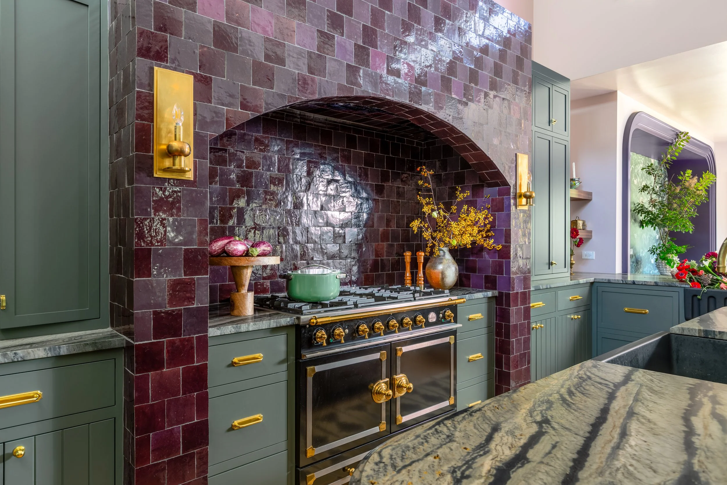 A kitchen with a bold dark red tile backsplash and green cabinetry accented with gold hardware. Features a large stovetop with a modern oven, and decorative elements including a vase with yellow flowers, a wooden bowl with onions, and plants near a w