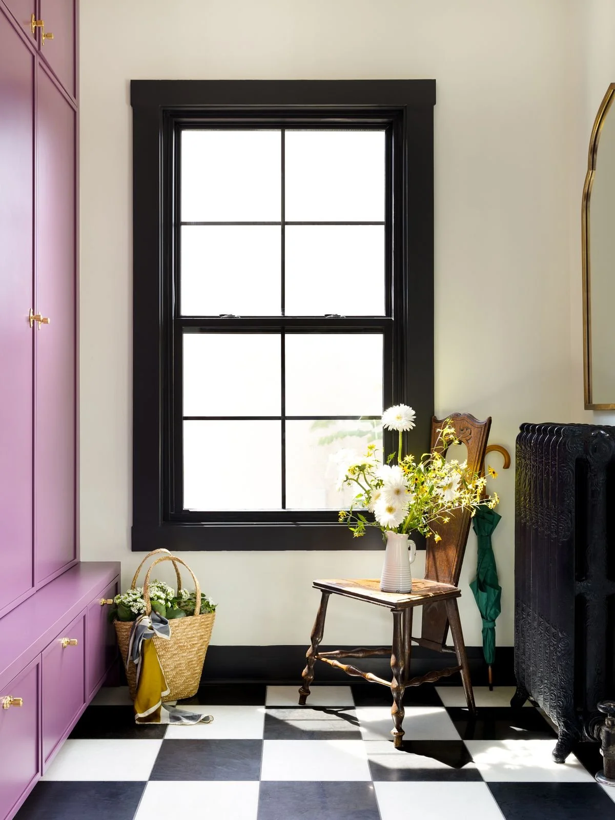 A room with a large black window, pink cabinets, a wicker basket with flowers, a wooden chair with a vase of white and yellow flowers, and black and white checkered floor.