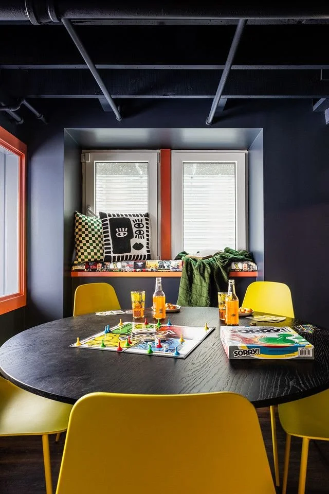 A round black table with yellow chairs, set up for a game of Sorry!, with six glasses of orange-colored drinks, and a game board in the center. Two large windows with shutters are in the background, above a window seat with patterned pillows and a gr
