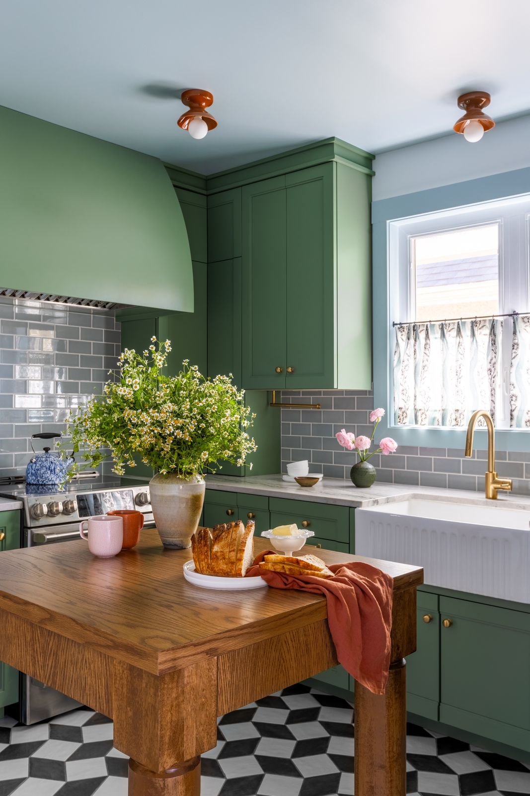 Kitchen with green cabinets, gray subway tile backsplash, window with floral curtains, wooden table with bread and drinks, flower vases, and black-and-white patterned floor.