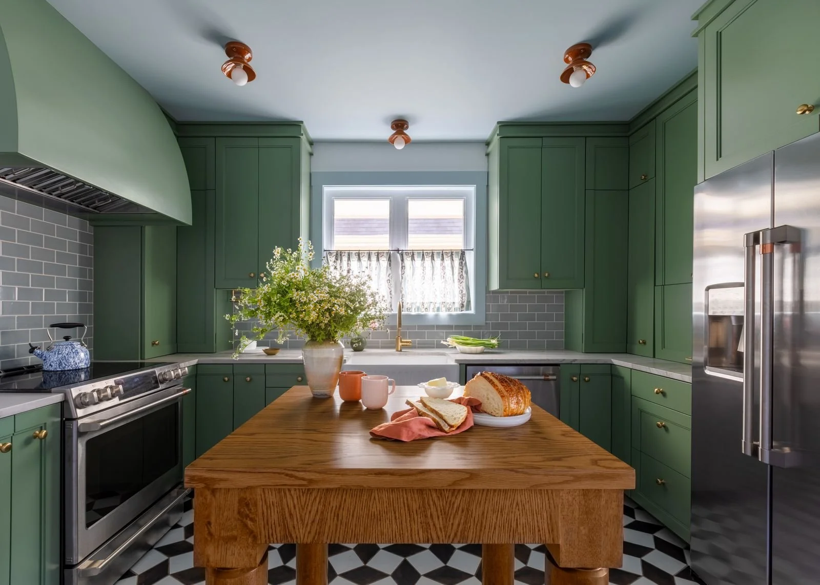 Kitchen with green cabinets, a wooden island with baked bread and breakfast items, gray subway tile backsplash, and a window with floral curtain