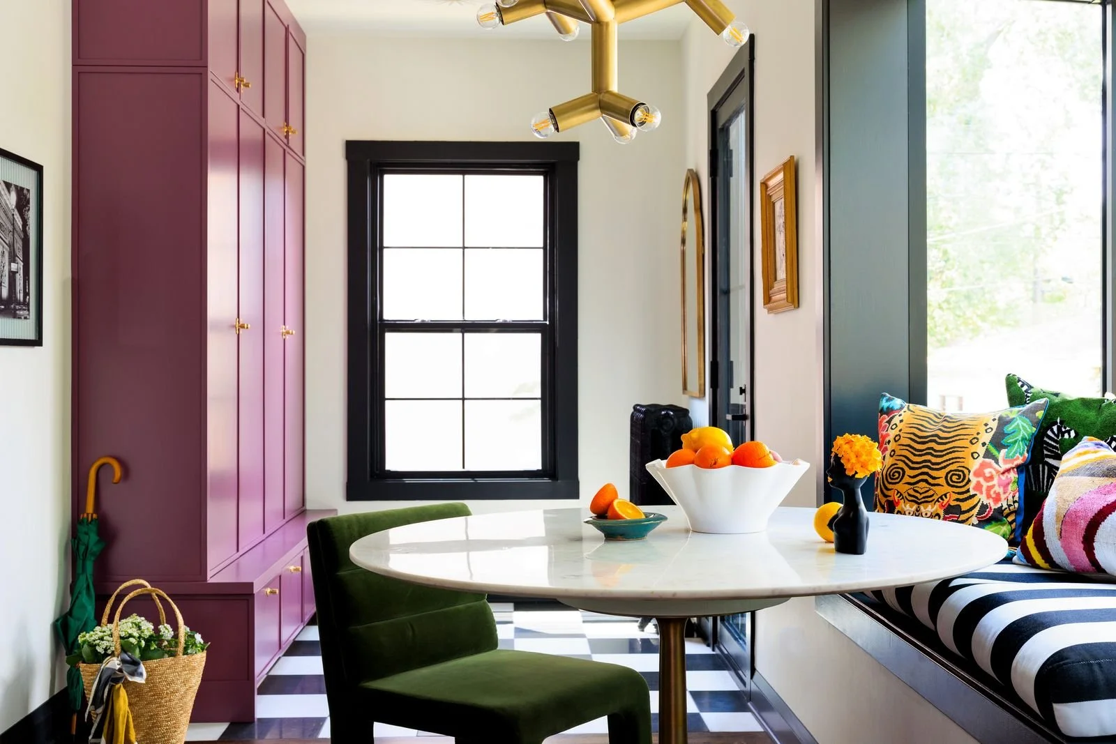 A colorful dining area with a white marble table, green velvet chair, built-in window seat with patterned pillows, black-framed window, and a modern gold chandelier. There are decorative items including a bowl of oranges and a small vase with yellow 