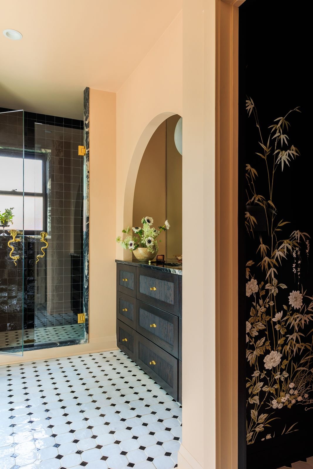 Interior view of a bathroom with black and white patterned floor, black dresser with brass knobs, a flower vase with white flowers, and a glass shower with black tiles and gold snake door handles.
