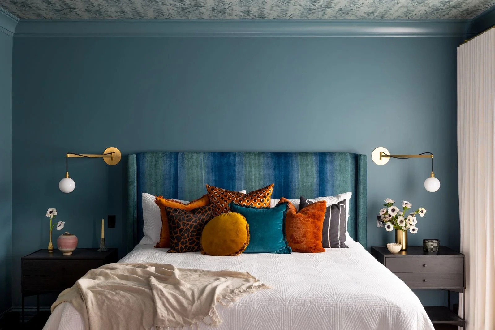 A modern bedroom with a blue upholstered headboard, colorful accent pillows, white bedding, two black nightstands with decorative items, wall-mounted lamps, and white curtains.