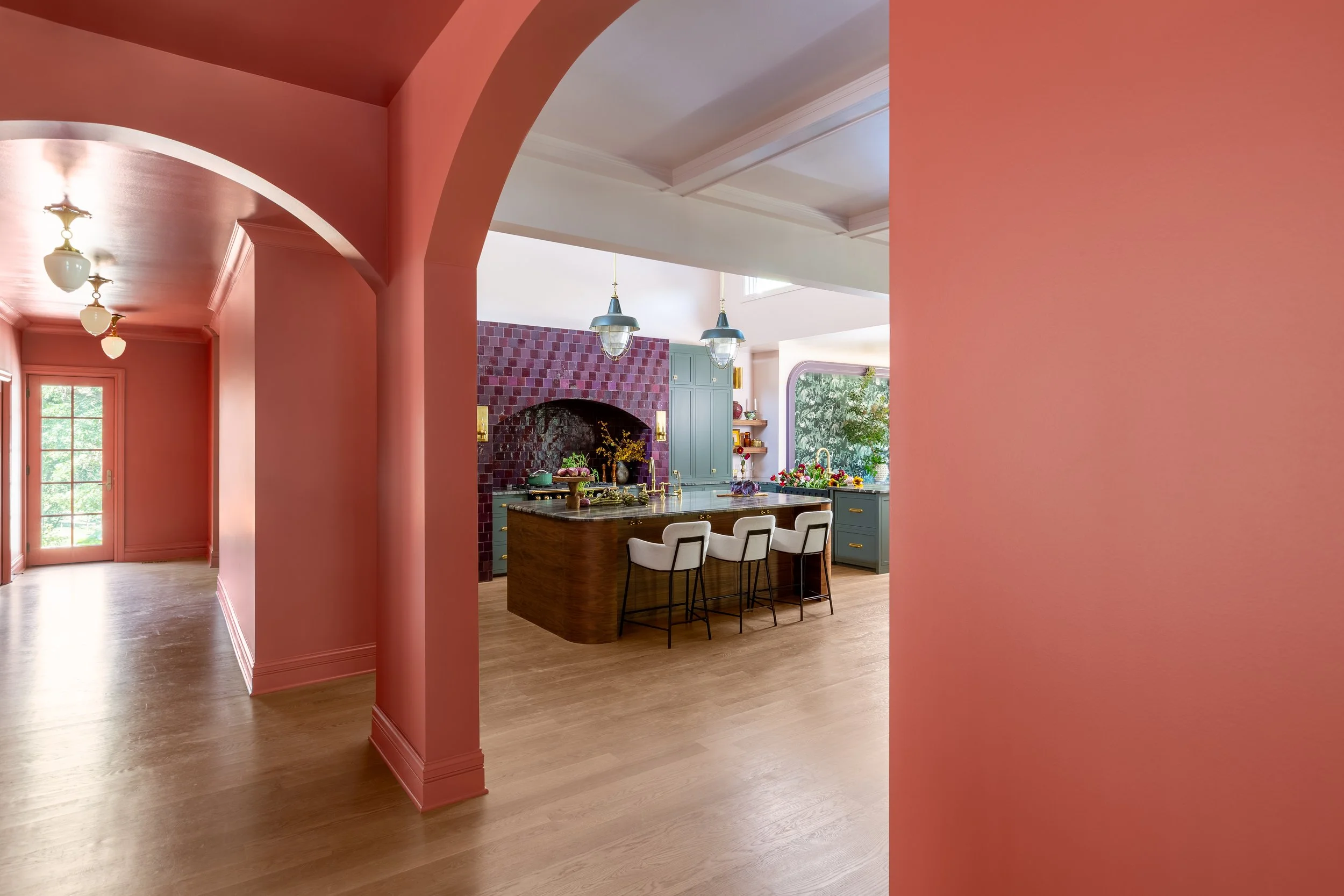 Open-concept kitchen and living space with pink walls, wooden floors, a kitchen island with white bar stools, blue cabinets, a brick purple backsplash, gold fixtures, hanging pendant lights, a large window with a view of greenery, and a door leading 