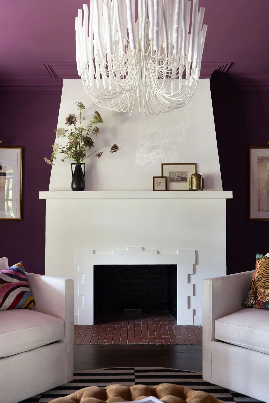 Living room with a white fireplace, purple walls, a modern chandelier, a black vase with dried flowers on the mantel, and frames on the shelf above the fireplace. Two white sofas with colorful pillows.